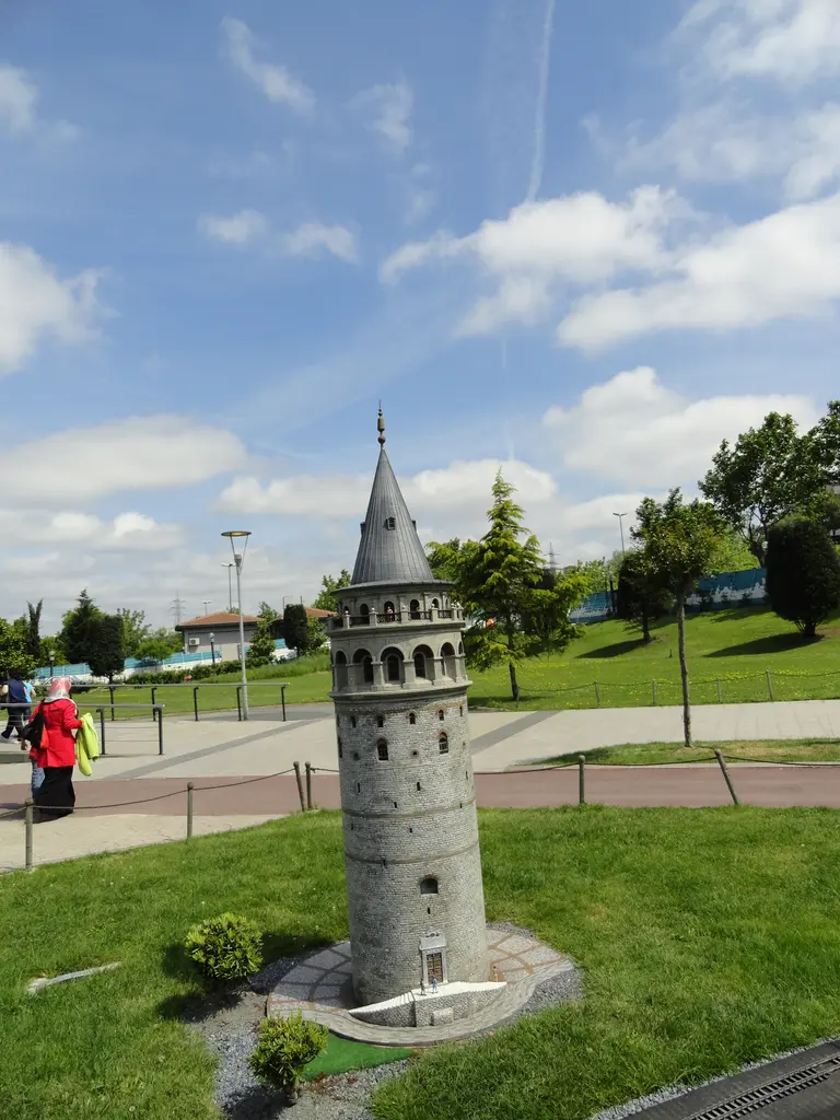 Miniatürk'te Galata Kulesi maketi; gri taş silindir gövde, konik külah, çimen üzerinde, arkada yollar, ağaçlar ve bulutlu gökyüzü.