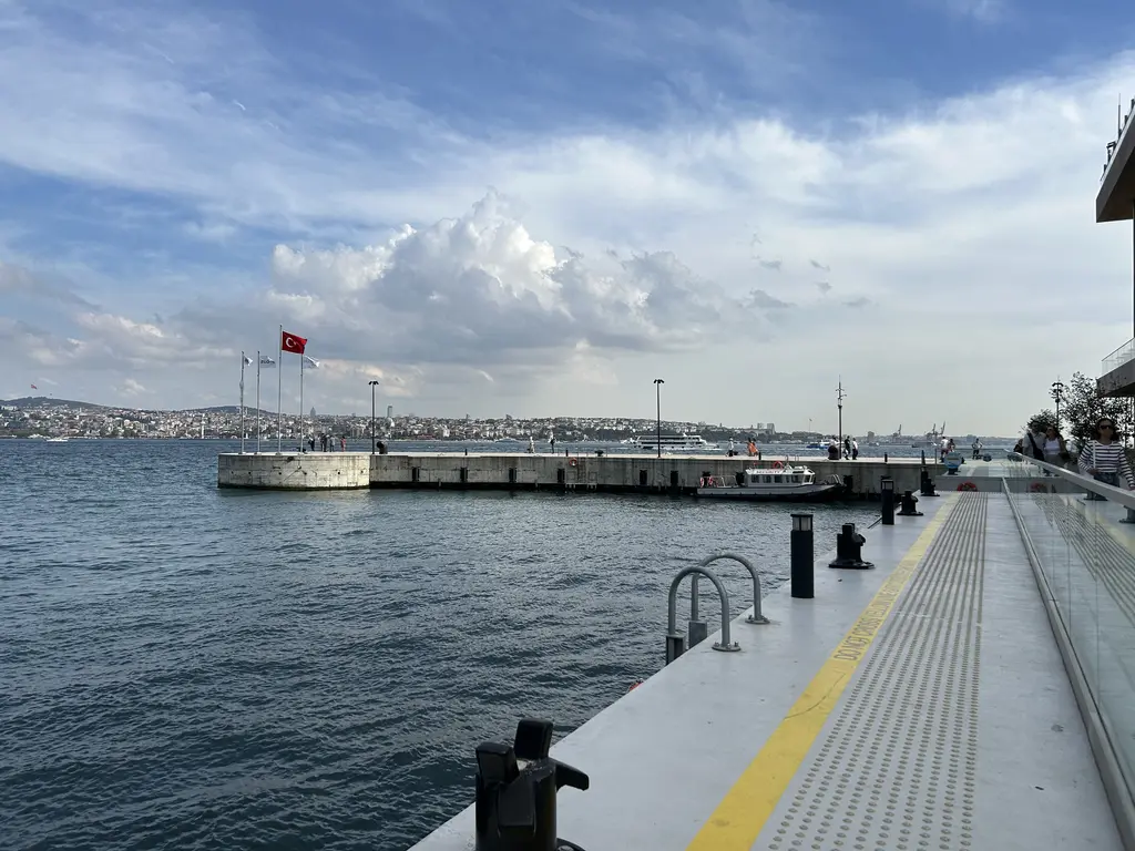 İstanbul Modern önündeki rıhtım; sarı çizgili iskele kenarı, Boğaz'ın dalgalı suyu ve uzakta karşı kıyı ve bulutlu gökyüzü.