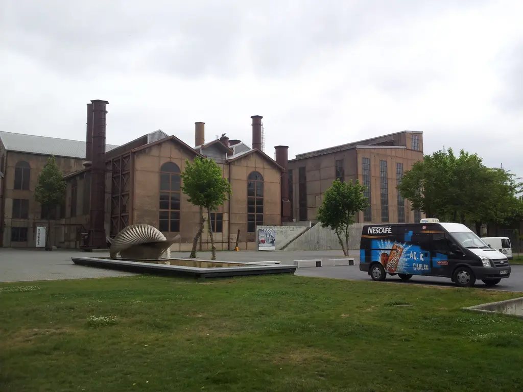 Santralistanbul'da makine dairesi; bacalı tuğla bina, önde çimen, ağaçlar, havuz kenarında metal heykel ve park etmiş araç.
