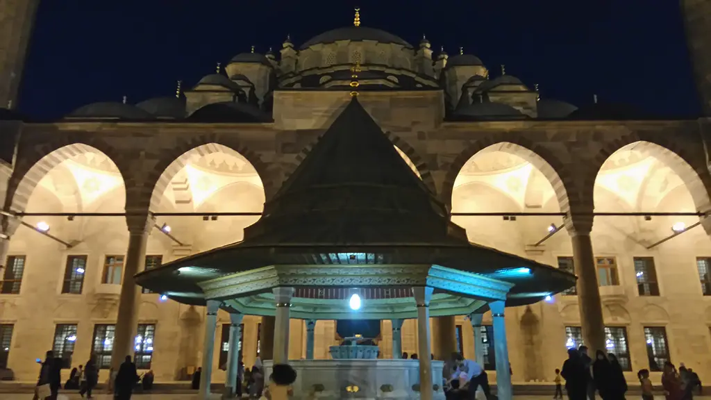 Fatih Camii'nin avlusu mavi saatte; lacivert gökyüzü altında amber ışıkla aydınlanan revak ile kubbeler, ortada turkuaz ışıklı şadırvan ve avluyu geçen siluetler.