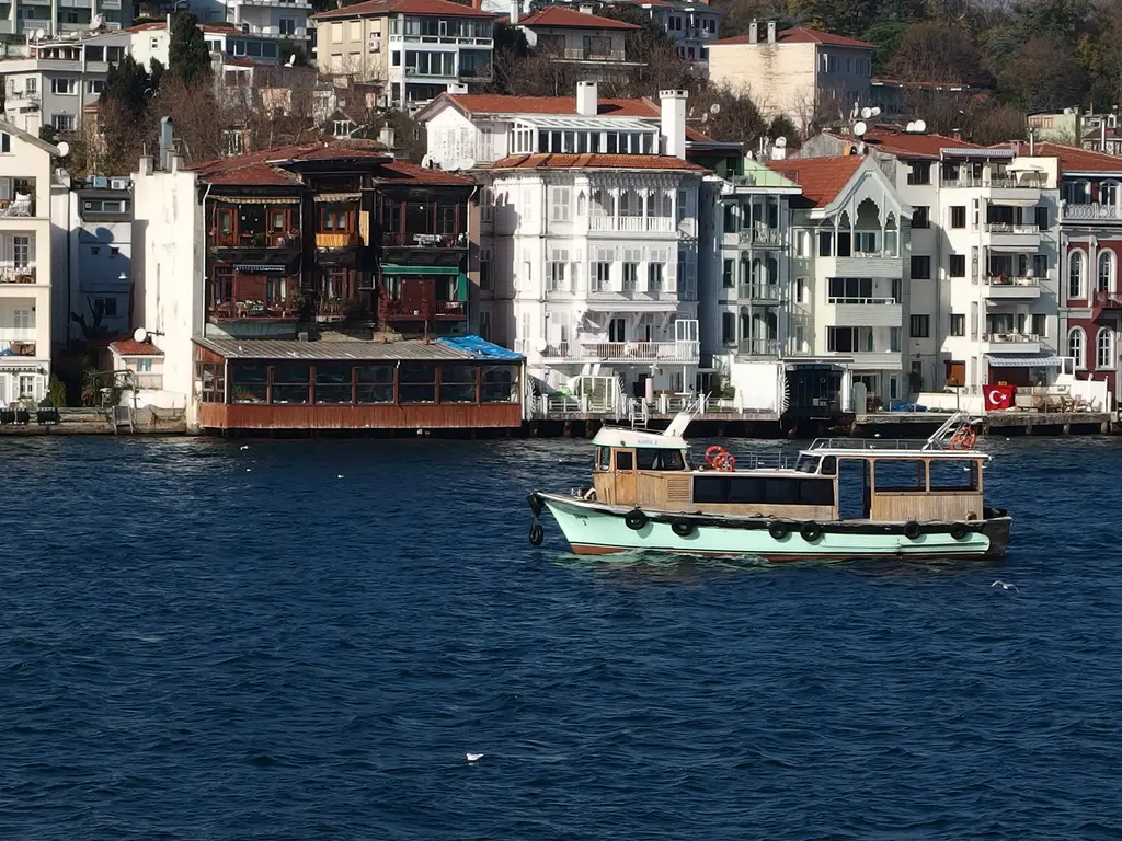 Yeniköy kıyısında koyu ahşap yalı ve beyaz komşuları; önde geçen yeşil teknecik ve arkada yamaca tırmanan evler.