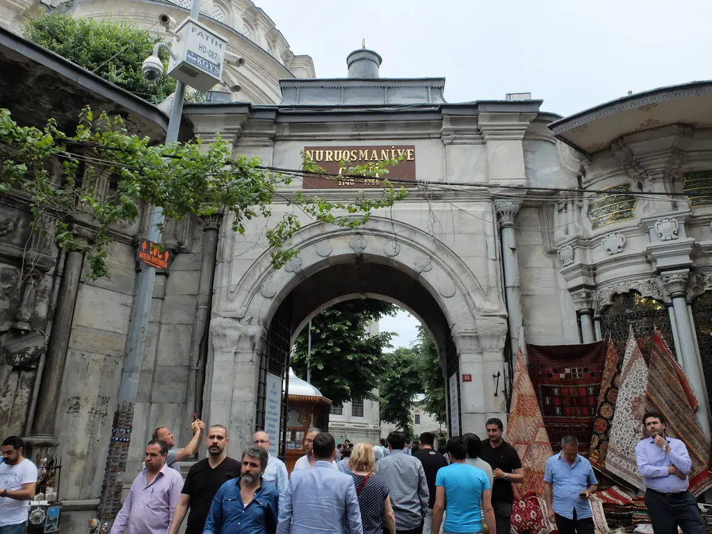 Nuruosmaniye Camii'nin çarşı sokağına açılan kemerli kapısı; üstünde caminin adını taşıyan tabela, kemerin ardında avlu ile minare, sağda asılı halılar.