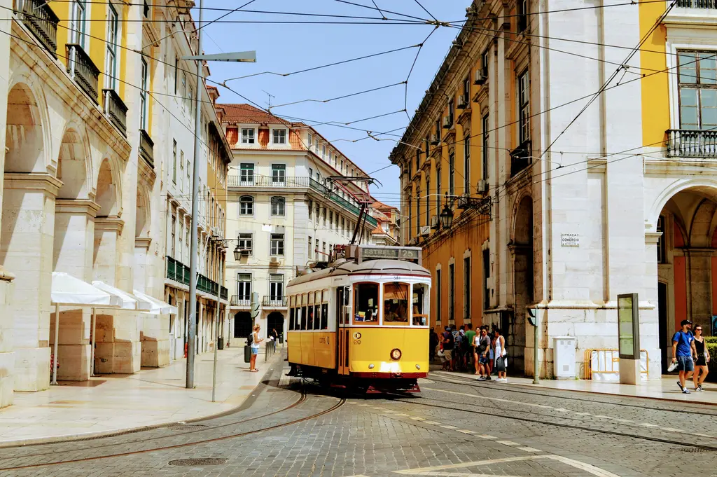 Lizbon'da tarihi bir sarı tramvayın Praça do Comércio yakınındaki taş binalar arasından geçişi; arnavut kaldırımı cadde ve yaz güneşi.