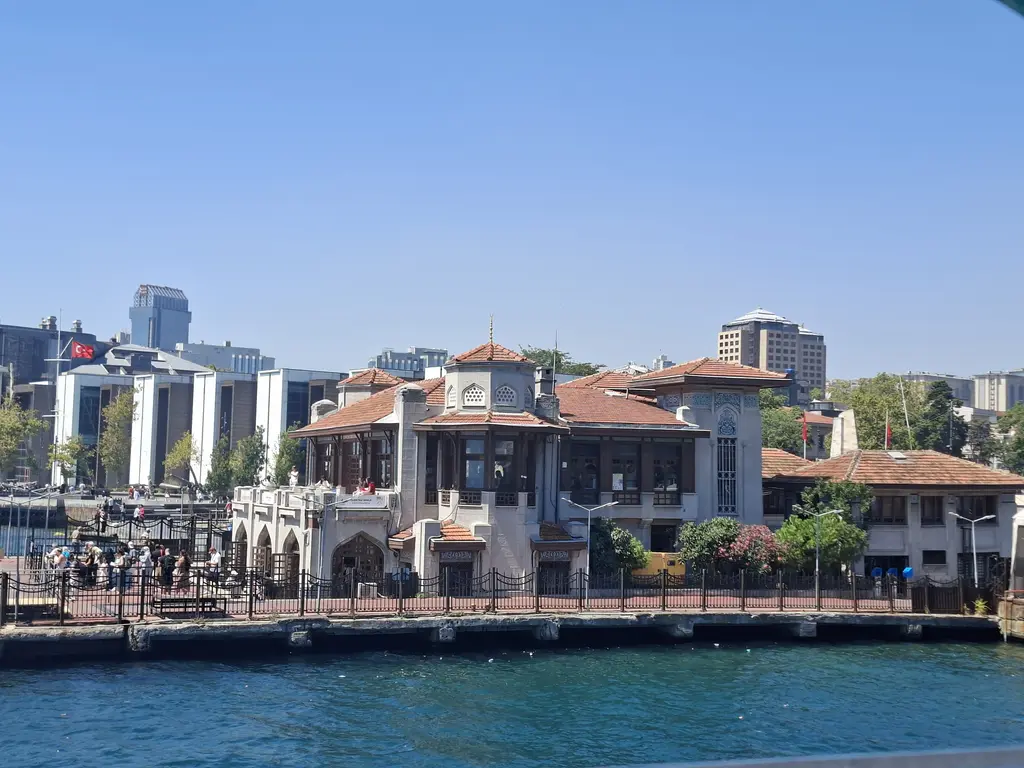 Beşiktaş iskelesi; kırmızı kiremitli çatılı iskele binası, önde rıhtım ve deniz, arkada Beşiktaş'ın binaları ve mavi gökyüzü.