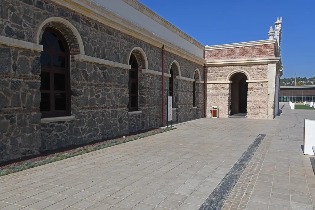 Feshane'nin yan cephesi; taş ve tuğla duvar, kemerli pencereler, köşede beyaz mermer kapı ve taş döşeli meydan.