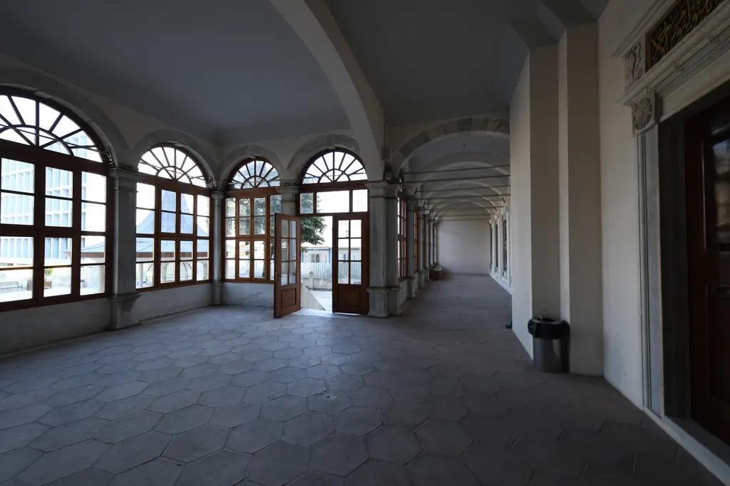 Nusretiye Camii'nin yan sofası; kemerli ahşap pencereler, uzun tonozlu koridor, altıgen taş döşeme ve açık kapıdan görünen dış mekân.