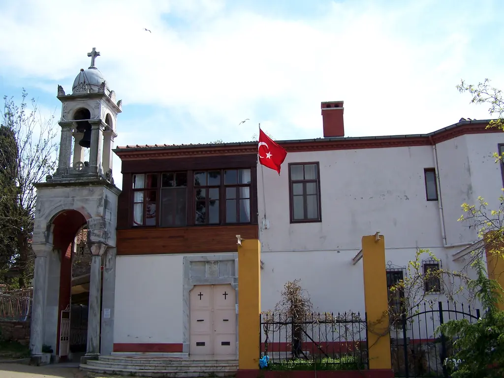 Büyükada'da Aya Yorgi Manastırı; kemerli çan kulesi, kırmızı çerçeveli ahşap çıkma, bayrak ve demir parmaklıklı bahçe.
