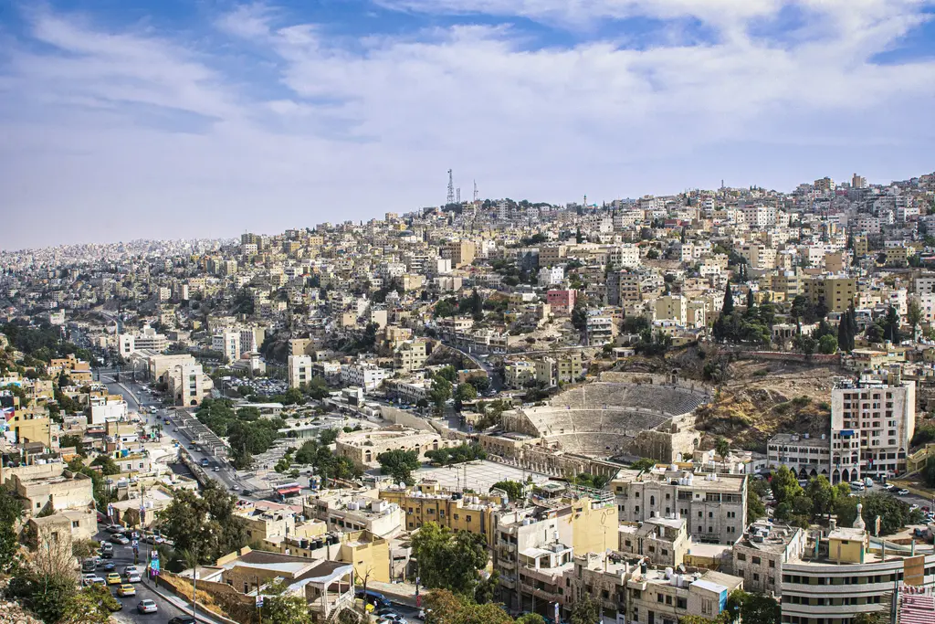 Amman'da tepelere yayılmış şehir manzarası; birbirine bitişik açık bej taş binalar, yamaçları izleyen sokaklar, ufuktaki anten kulesi ve puslu gökyüzü.