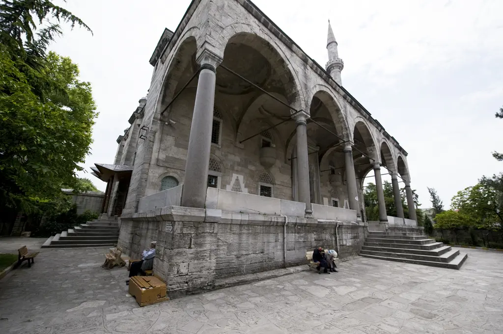Caminin yan cephesi; yüksek subasman üzerinde beş sıra pencere, üstte yarım kubbeler ve ağırlık kuleleri, önde ağaçlar.