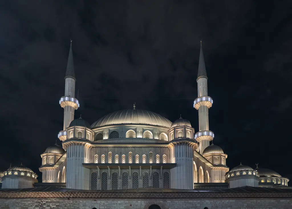 Gece ışıklandırılmış Taksim Camii; beyaz taş cephe, katmanlı kubbeler ve iki ince minare, arkada koyu gökyüzü.
