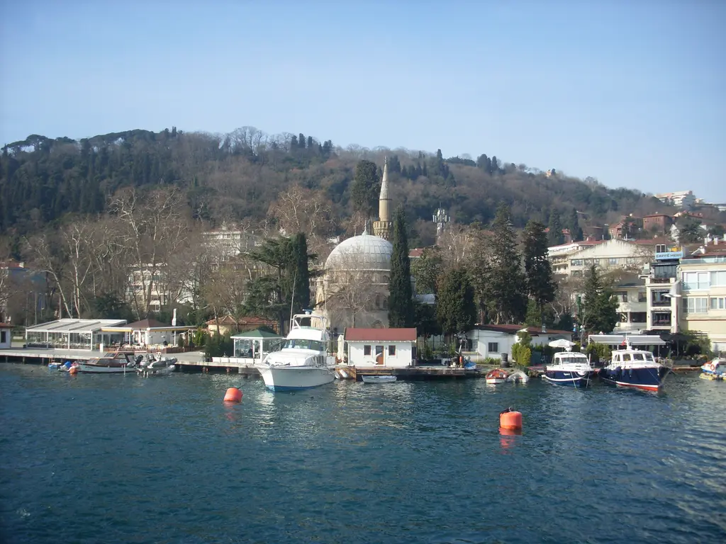 Bebek Koyu; demirli tekneler ve şamandıralar, kıyıda cami ve minare, arkada ağaçlık yamaç ve evler.