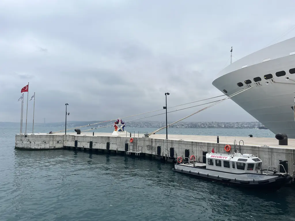 Galataport'ta iskele; sağda kruvaziyer gemisinin beyaz pruvası, önde küçük kılavuz teknesi, solda bayraklar ve Boğaz.
