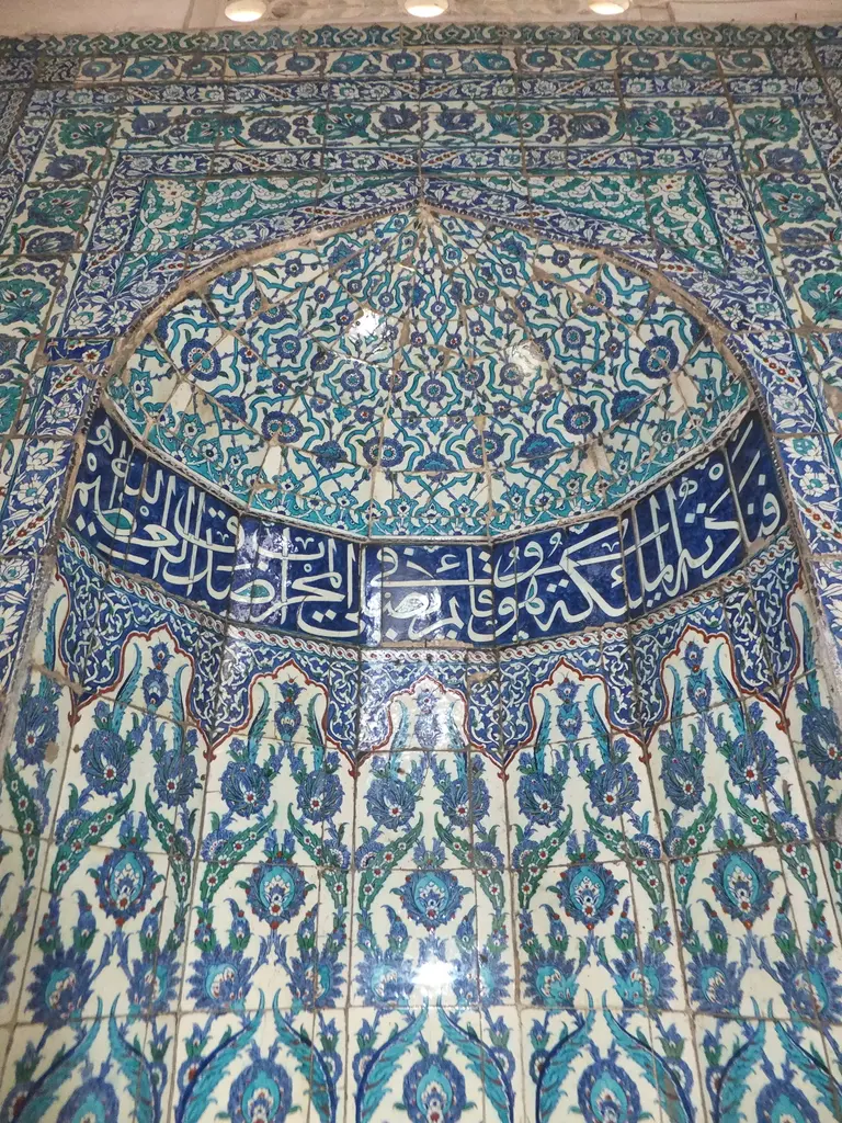 Çinili Camii'nde pencere alınlığı; kemerli çini pano, beyaz harfli lacivert yazı şeridi ve çiçek desenleri.
