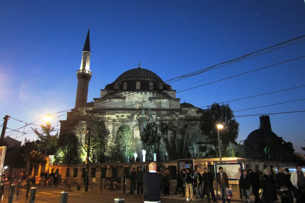 Akşam Gazi Atik Ali Paşa Camii; lacivert gökyüzü, ışıklı minare, avlu duvarı önünde tramvay durağı, sokak lambaları ve yayalar.