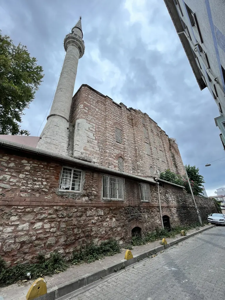Gül Camii'ne eğimli sokaktan bakış; kaldırım hizasında küçük kemerli açıklıkları olan yüksek taş bodrum, üstünde gövde ve solda minare.