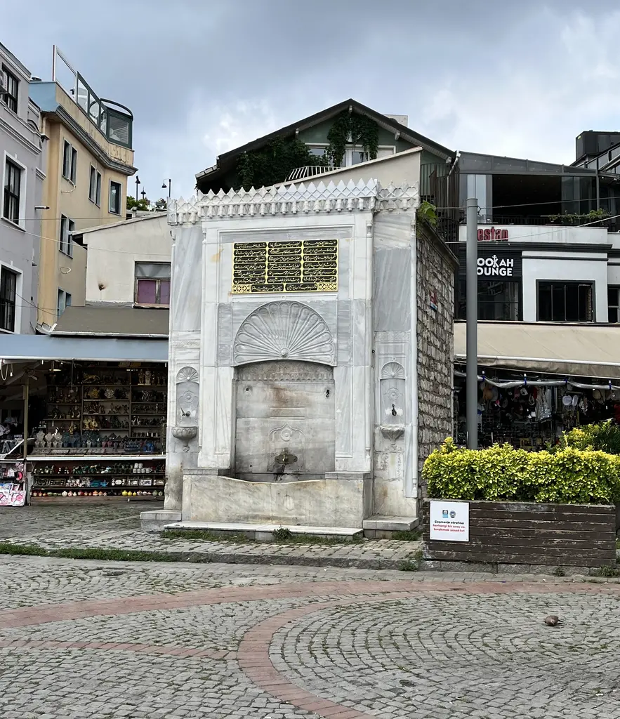 Ortaköy meydanında Damat İbrahim Paşa Çeşmesi; beyaz mermer cephe, kemerli niş, çevresinde çiçekler ve arkada evler.