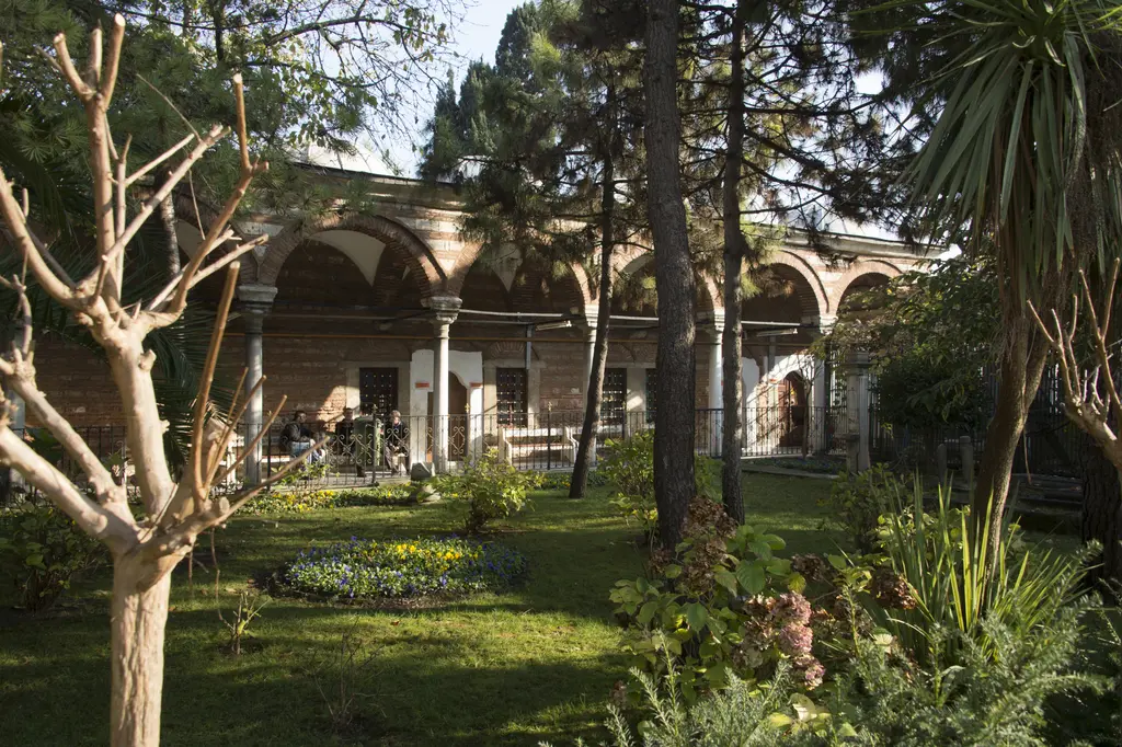 Mihrişah Valide Sultan İmareti'nin bahçesi; çimenler, çiçek tarhı, palmiye ve ağaçlar, arkada sütunlu kemerli revaklar.