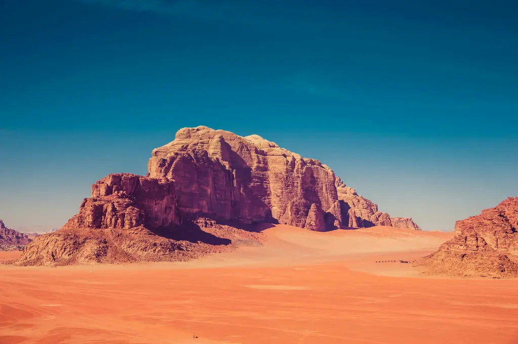 Wadi Rum'da kızıl kum çölü; ortada dik yüzlü geniş bir kumtaşı kütlesi, önünde dalgalı kum yüzeyi ve üstünde bulutsuz mavi gökyüzü.