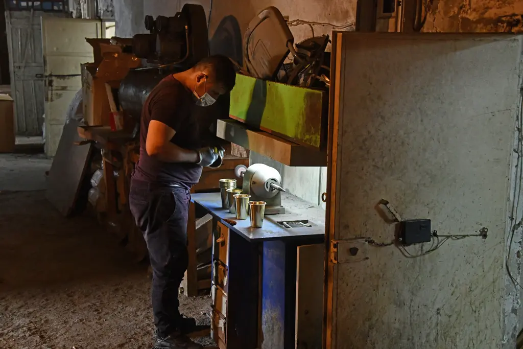 Büyük Valide Han'da loş bir atölye; maskeli bir usta polisaj motorunda metal bardak parlatıyor, tezgâhta sıralanmış bardaklar ve sağda eski demir kapı.