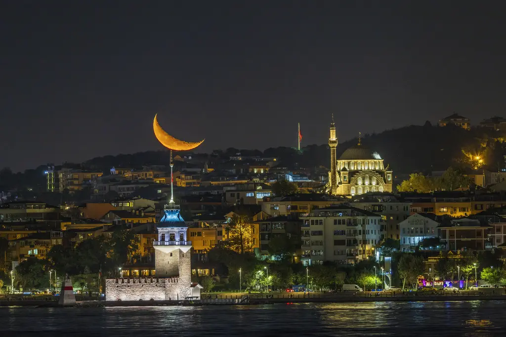 Gece Kız Kulesi; ışıklı kule, arkasında Üsküdar'ın aydınlatılmış camileri ve evleri, gökyüzünde hilal.