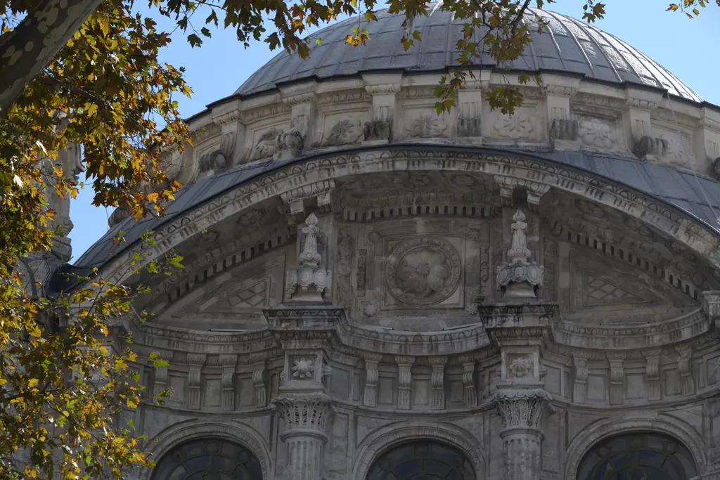 Ortaköy Camii'nin kubbesi; ağaç dallarının arasından görülen kurşun kubbe, kemerli kasnak ve taş süslemeler.