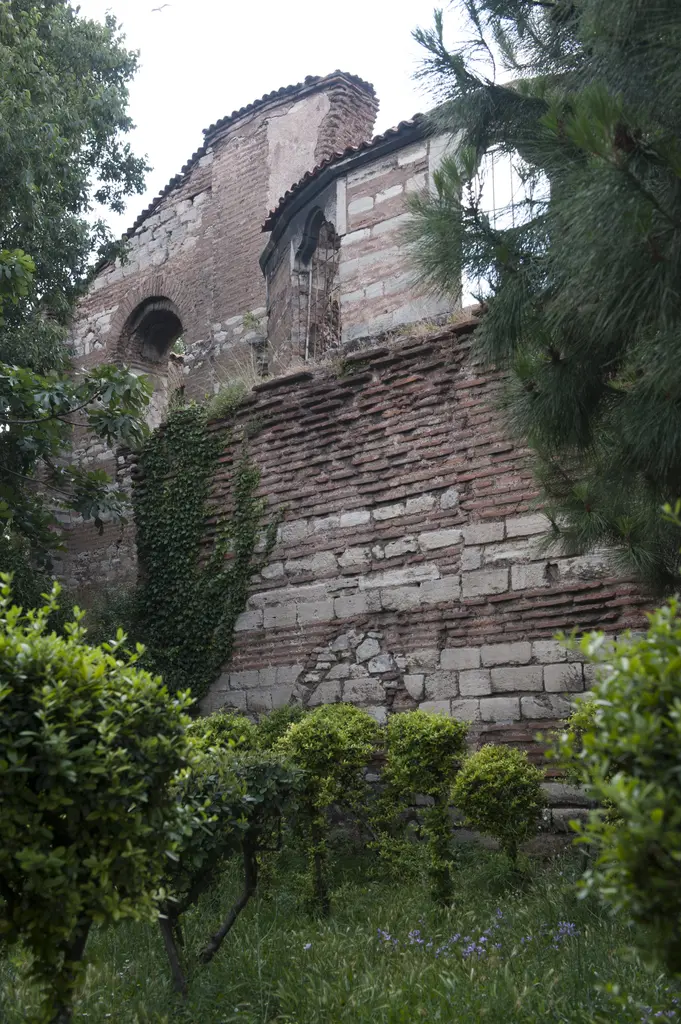 İmrahor İlyas Bey Camii'nin apsisi bahçeden; altta sarmaşık kaplı taş tuğla gövde, üstte kemerli pencereli kasnak ve kiremit saçak, önde çam ile yeşillik.