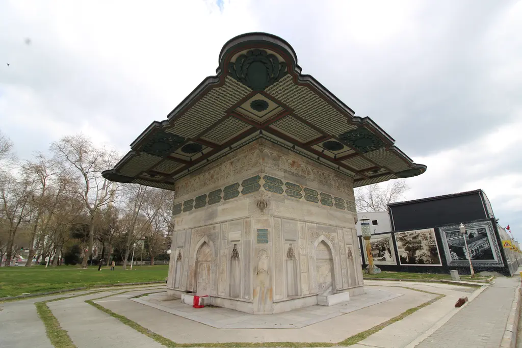 Tophane Çeşmesi; geniş saçak ve kare gövde, çevresinde ağaçlar ve çimen, önde meydanın taş döşemesi ve yürüyüş yolları.
