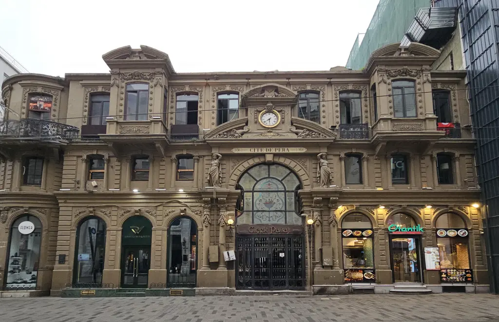 İstiklal Caddesi'nde Çiçek Pasajı'nın ön cephesi; üç katlı süslü taş yapı, ortada renkli camlı kemerli giriş, üstünde saat ve zemin katta dükkânlar.