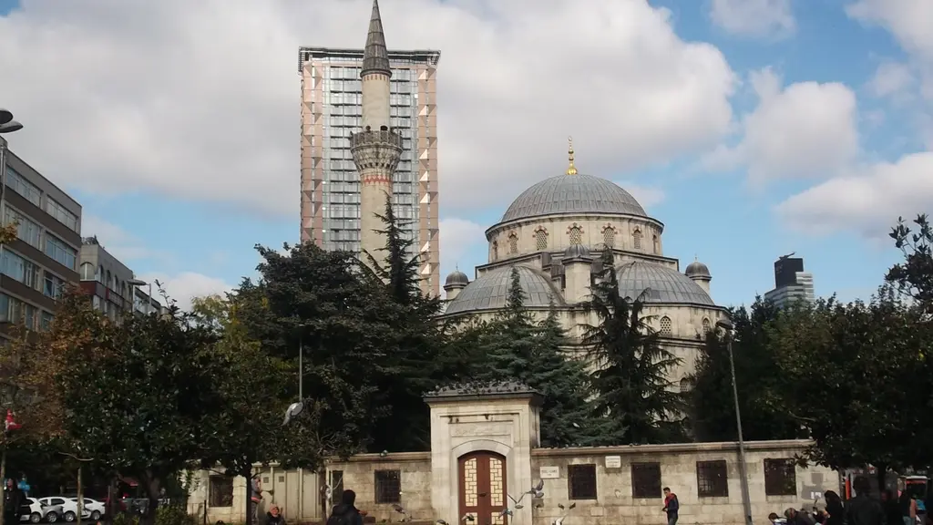 Şişli Camii'ne meydandan geniş bakış; uzun avlu duvarı, ağaçlar arasında kurşun kubbeler, minare ve arkada yüksek bir apartman bloğu.