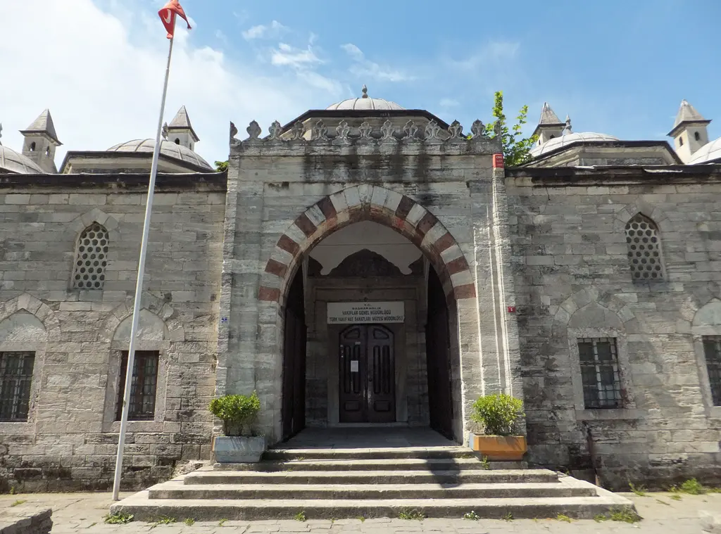Medresenin girişi; kemerli taç kapı, iki yanda kemerli pencereler, önünde bayraklar, saksılar ve taş döşeli meydan.