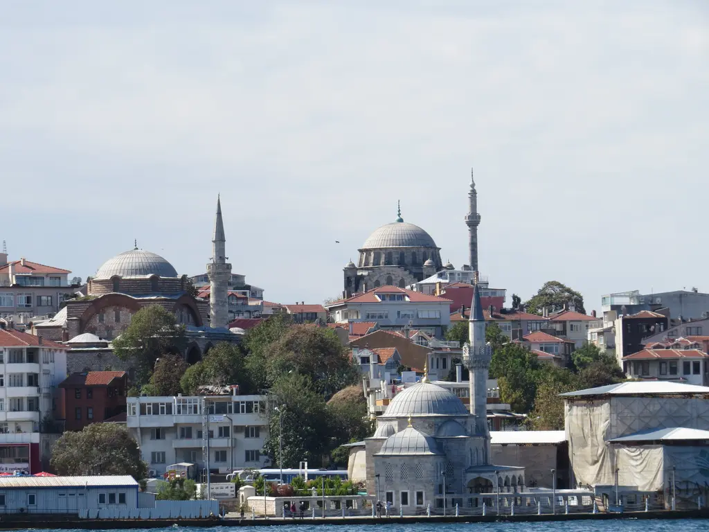Şemsi Paşa Camii; kıyıda kubbe ve minare, arkasında yamaçta iki büyük kubbeli cami ve ağaçlar.