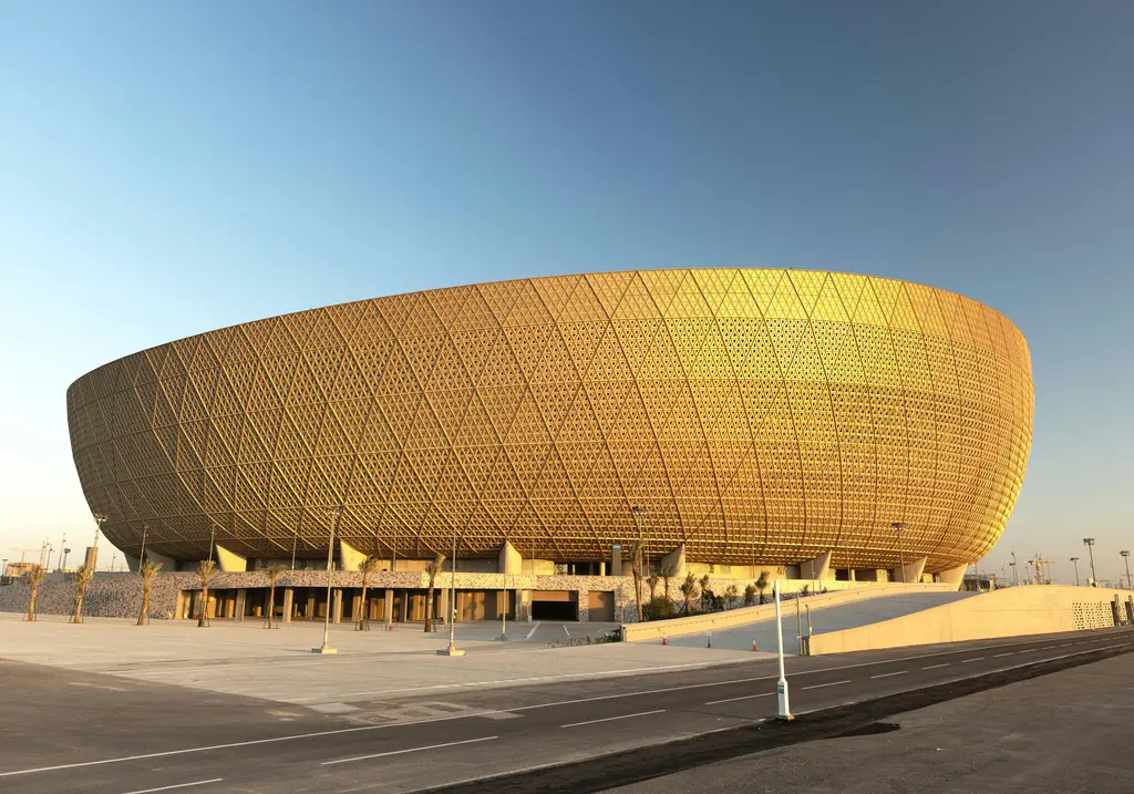 Lusail'de altın rengi stadyum; delikli metal cephesiyle kâse biçimli gövdesi, çevresindeki geniş meydan ve boş yollar.