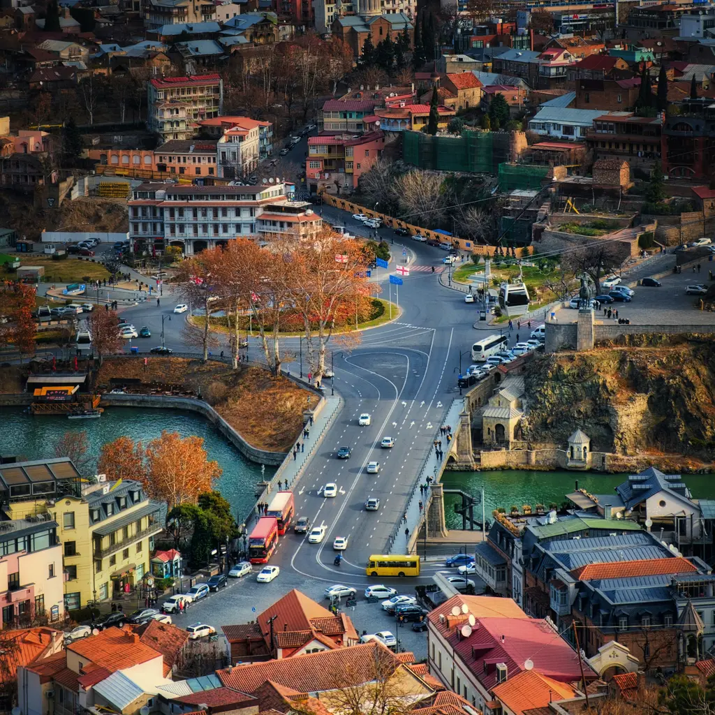 Tiflis'te Mtkvari Nehri'ne bakan sarp kayalığın tepesinde duran Metekhi Kilisesi; tuğla cephesi, koyu konik kubbesi ve yanındaki servi ağaçları.
