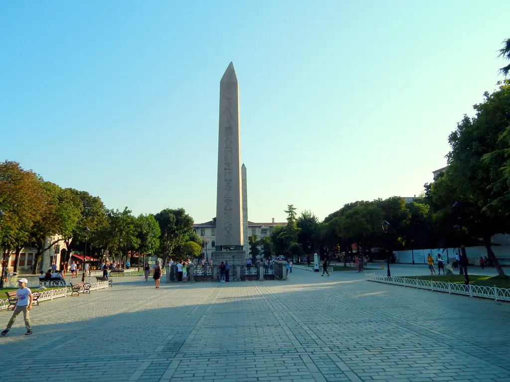 Güneşli bir günde Sultanahmet Meydanı; uzun taş döşeli yol, iki yanda ağaçlar ve yolun ortasında yükselen Dikilitaş.