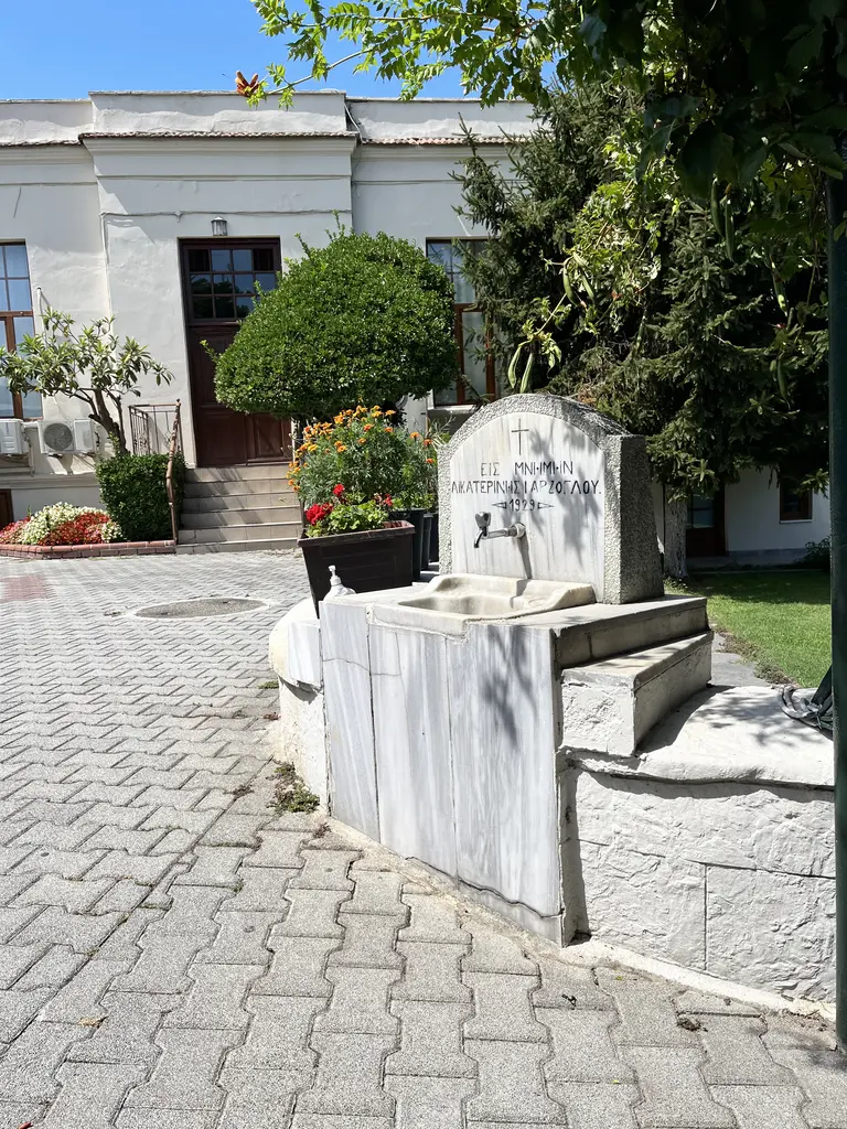 Avluda mermer bir anma çeşmesi; üstünde Rumca "Eis mnimin" diye başlayan yazıt, önünde yalak ve arkasında basamaklı ek bina ile çiçekler.