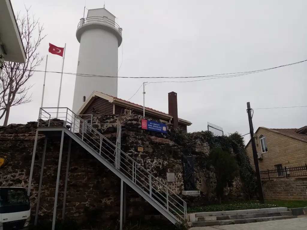 Anadolu Feneri; beyaz silindir fener kulesi, yanında bayrak direği, taş duvar ve merdivenler.