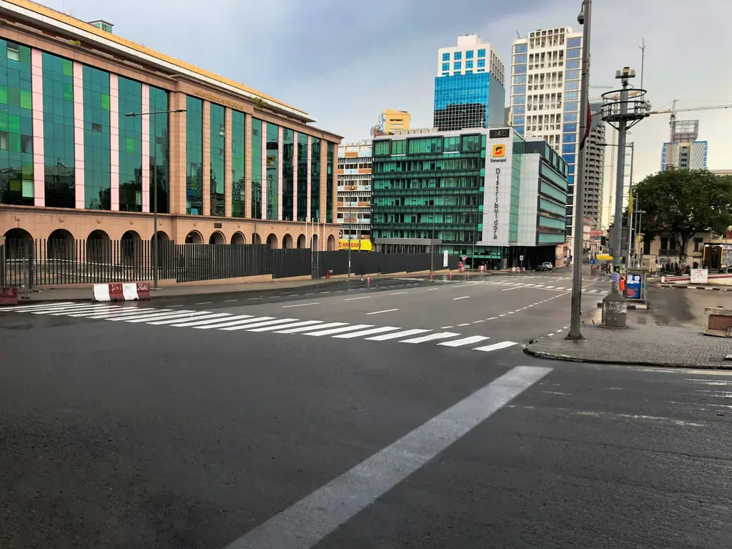 Luanda'da yağmur sonrası ıslak, ıssız bir kavşak; modern cam cepheli ofis binaları, gökdelenler ve yeni boyanmış yaya geçidi.