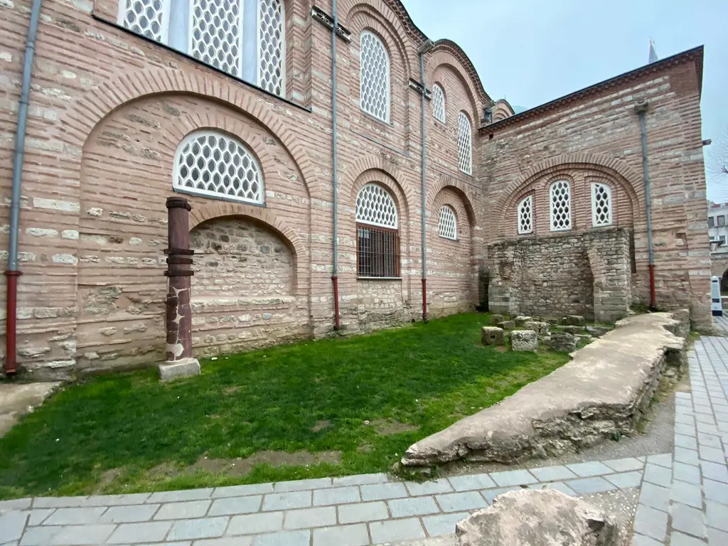 Zeyrek Camii'nin yan cephesi; kemerli kafes pencereler, tuğla ve taş sıralarıyla örülü duvar, önünde çim şerit ile dağınık duran işlenmiş taş parçaları.