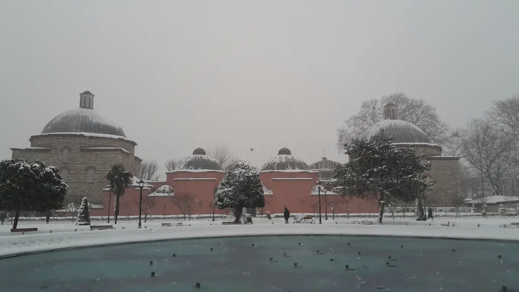 Kar yağarken hamam; beyazla kaplı kubbeler, pembe duvarlar, önünde donmuş havuz ve karla örtülü ağaçlar.