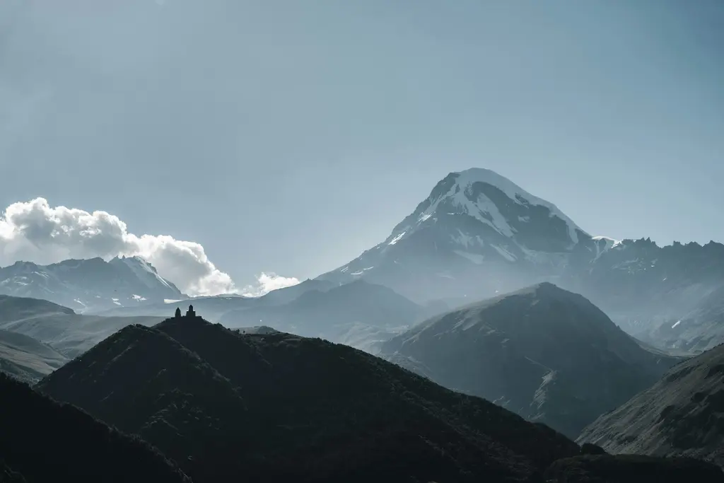 Kazbegi bölgesinde yeşil vadiler ve arkada karla kaplı sivri dağ zirveleri; gökyüzünde dağınık bulutlar.