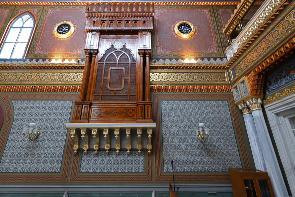 Yıldız Hamidiye Camii'nde duvara oturan ahşap mahfil; oymalı korkuluk, çini desenli duvar, yuvarlak levhalar ve aplikler.