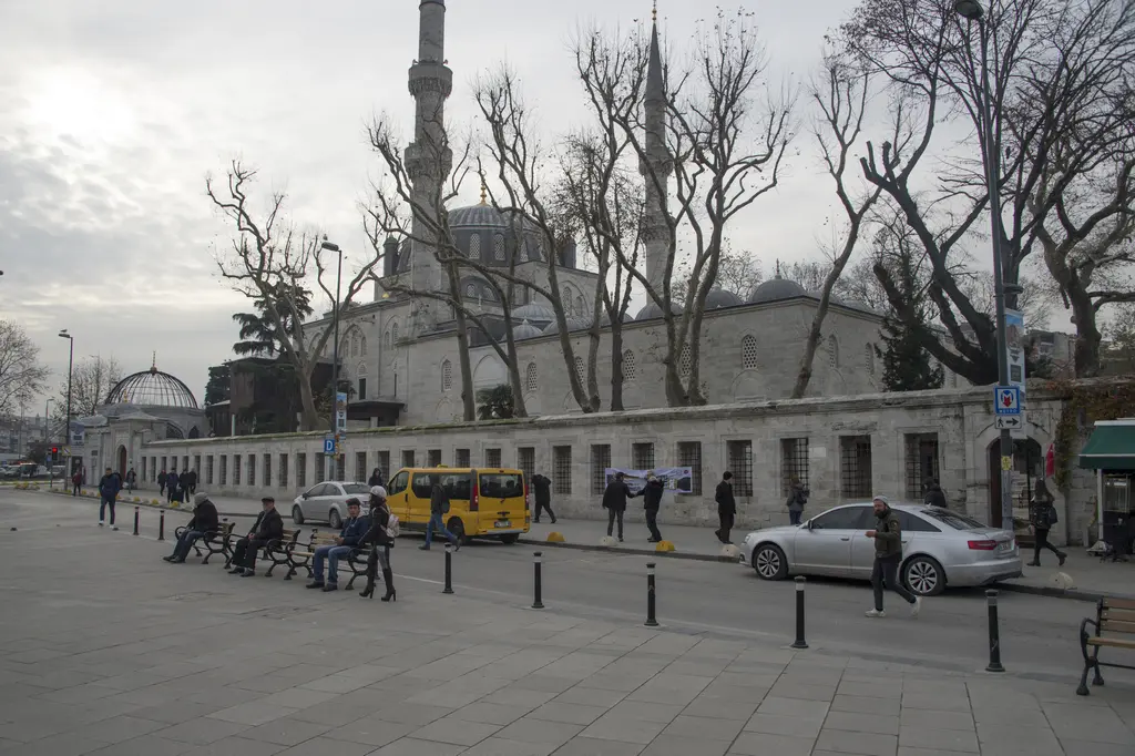Yeni Valide Camii'nin dış avlu duvarı; uzun taş duvar, önünde kaldırım, banklar ve arkada minareler.
