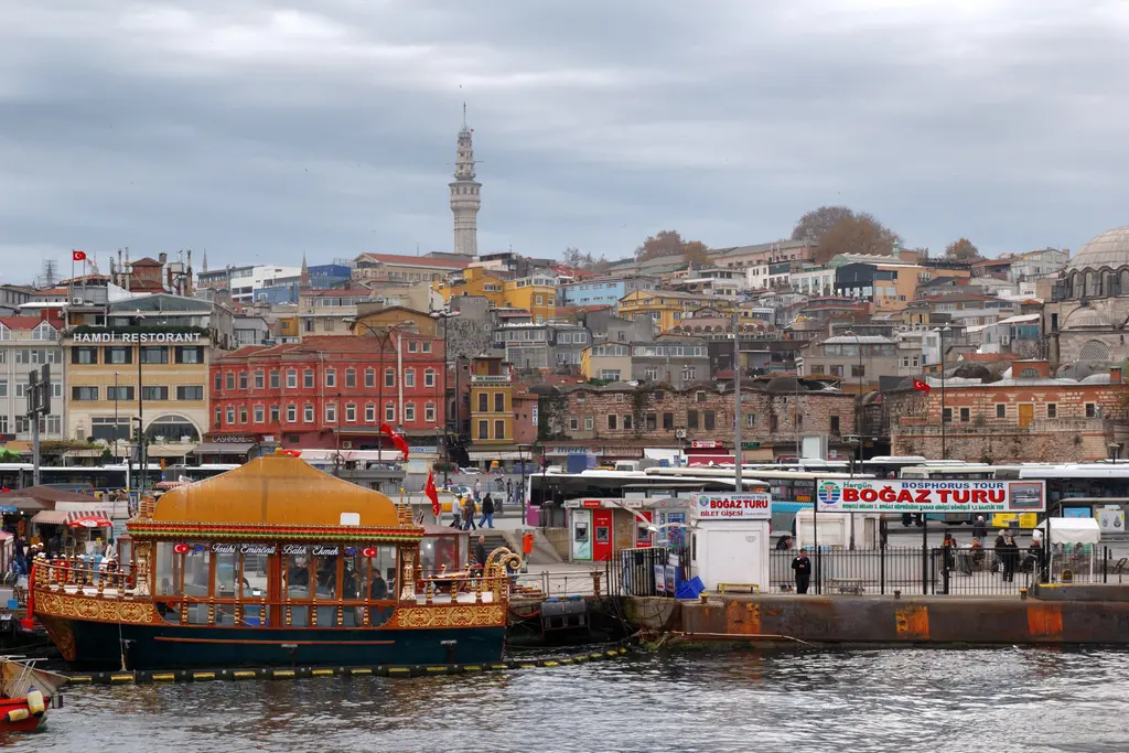 Eminönü kıyısı; rıhtıma bağlı turuncu çatılı tekne, Boğaz turu gişeleri, kıyıdaki binalar ve tepede Beyazıt Kulesi.