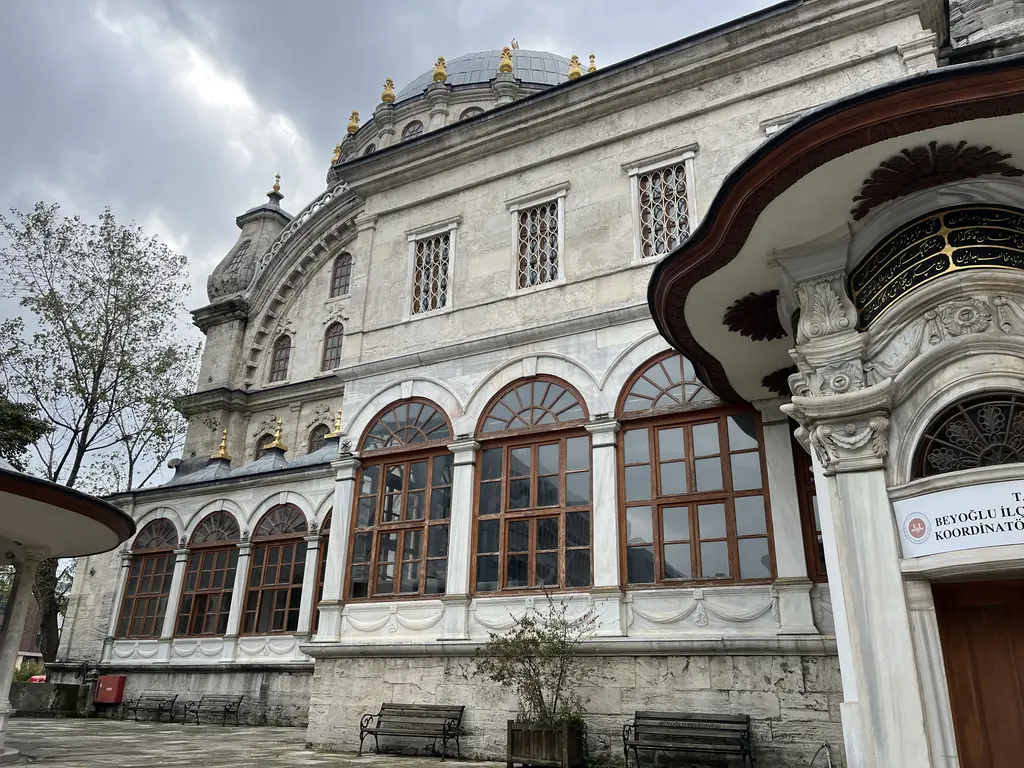 Nusretiye Camii'nin yan cephesi; kemerli büyük pencereler, mermer duvarlar, sağda dalgalı saçaklı sebil ve önde ağaçlar.