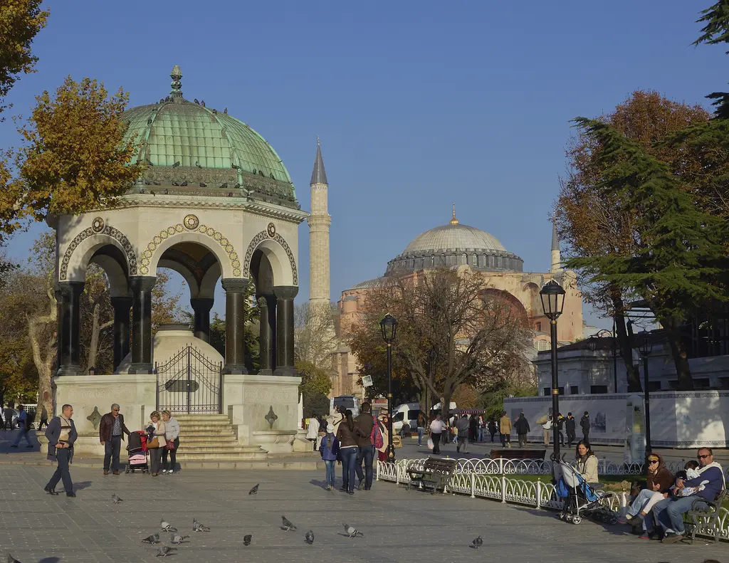 Sultanahmet Meydanı'nda Alman Çeşmesi; önde güvercinler ve banklar, ortada çeşme, arkada Ayasofya'nın kubbesi ve sonbahar ağaçları.