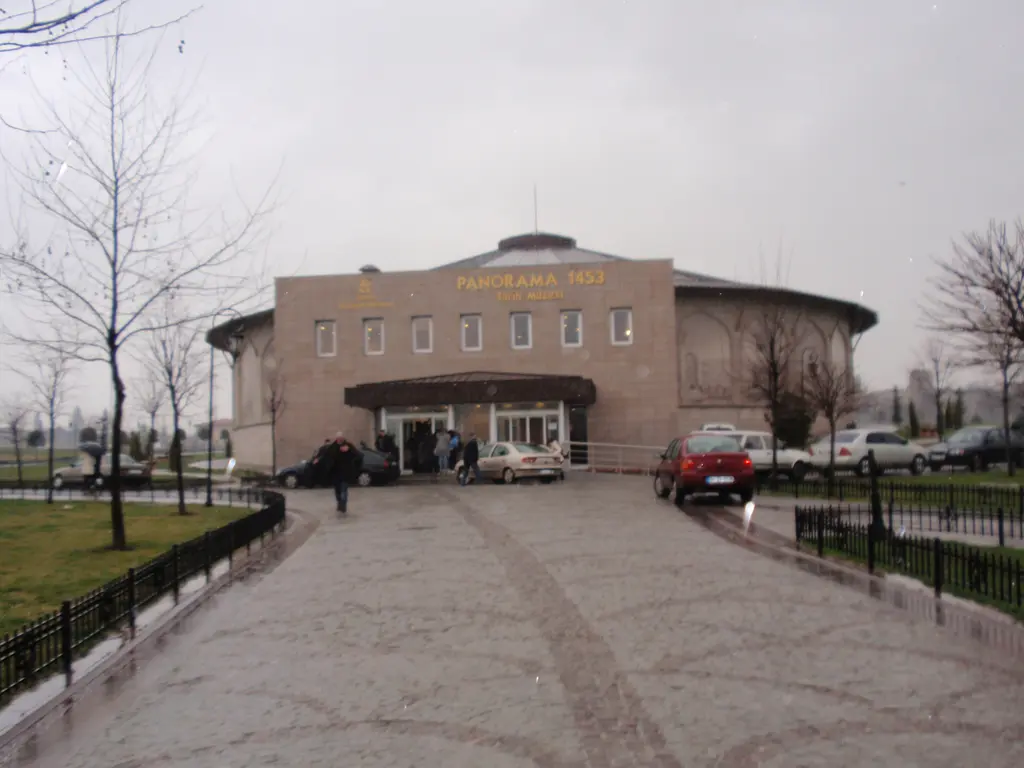 Yağmurlu bir günde müzenin giriş cephesi; alçak beton bina, cam kapılar, önünde ıslak meydan, demir parmaklık ve park eden araçlar.