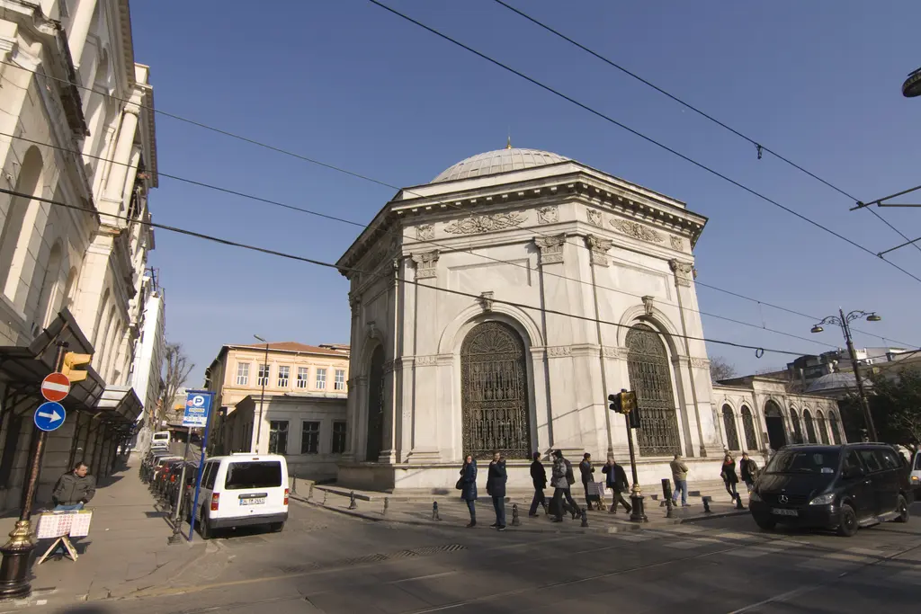 Divanyolu'ndan II. Mahmud Türbesi'ne geniş açılı bakış; sekizgen mermer türbe, yanında avlu duvarı, tramvay telleri, park etmiş araçlar ve yayalar.