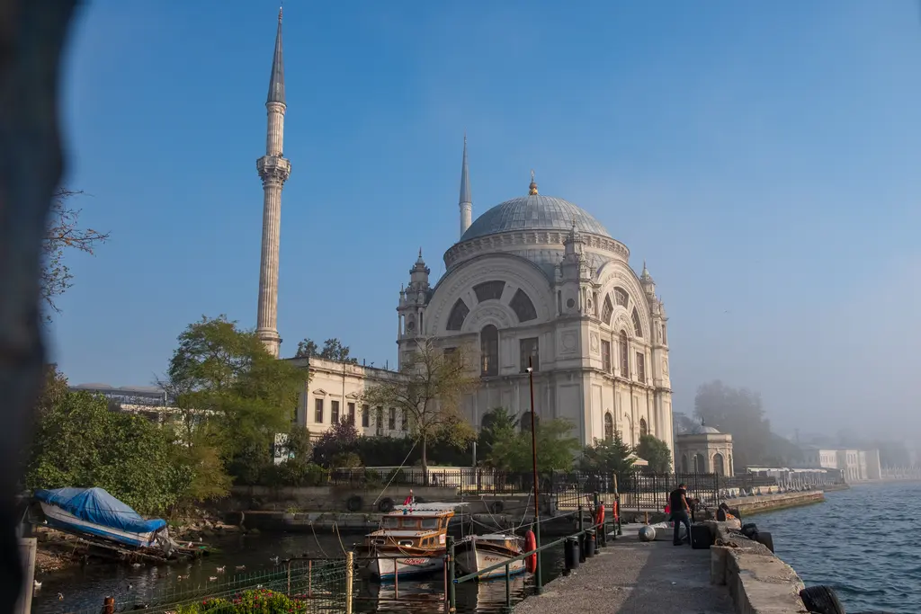 Kıyıdan Nusretiye Camii; sisli bir sabah, kubbe ve ince minare, önde ağaçlar, rıhtım boyunca yürüyen insanlar ve bağlı tekneler.