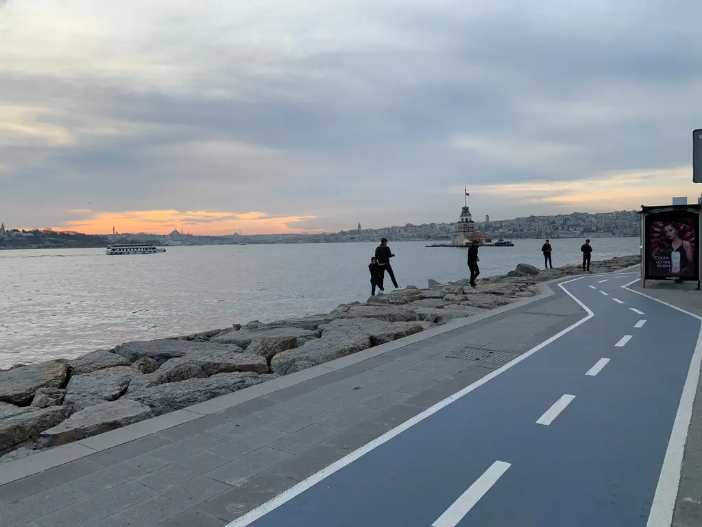Salacak'ta sahil yolu; bisiklet yolu, kıyıdaki taş dolgu, yürüyen insanlar ve uzakta Kız Kulesi.
