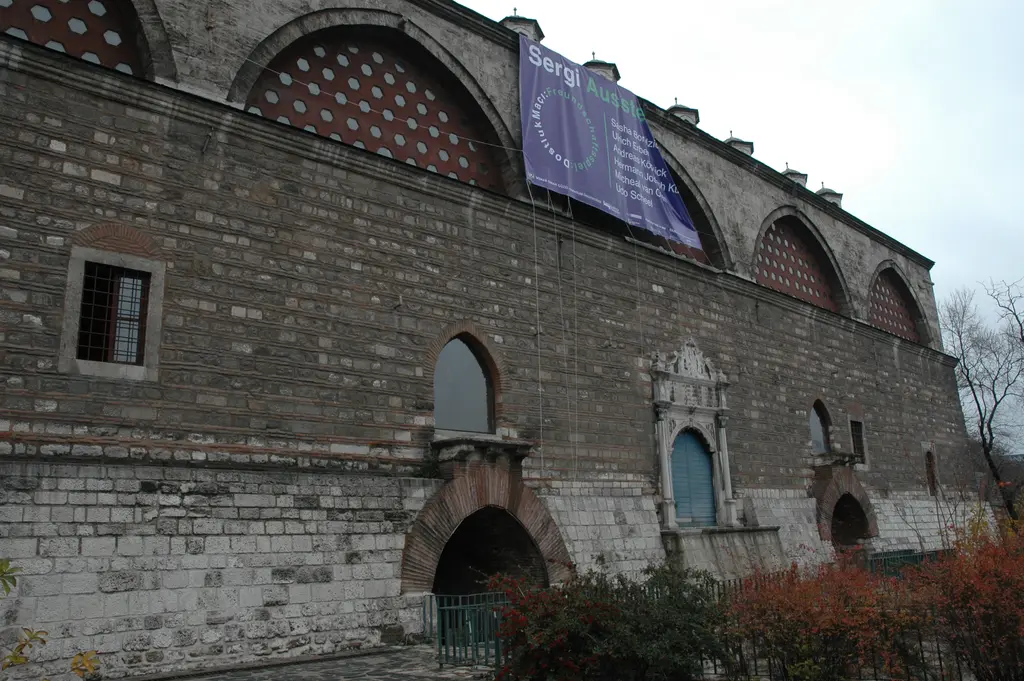 Tophane-i Âmire'nin uzun taş cephesi; üstte delikli kemerler, duvarda asılı bir sergi afişi ve önde kırmızı yapraklı çalılar.